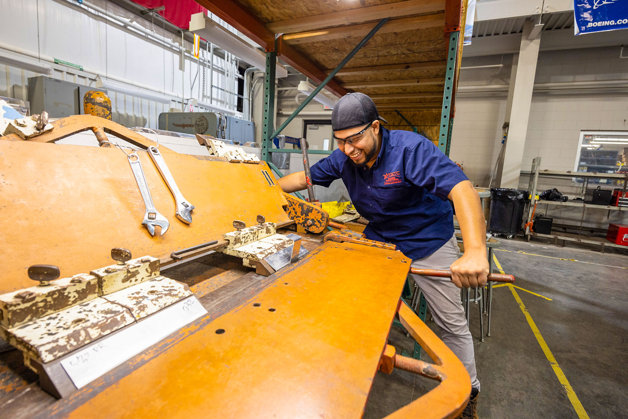 Student wearing safety glasses operates a large orange metal shear, pulling a long lever to cut sheet metal in a workshop with tools and equipment nearby.